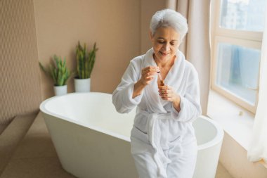 Cilde doğal damla kozmetik yağı süren bir kadın. Yetişkin yaşlı bir kadın günlük güzellik rutini yapan pipetli anti-yaş serumu uyguluyor. Yaşlı kadın yüz bakımı yaptırıyor.