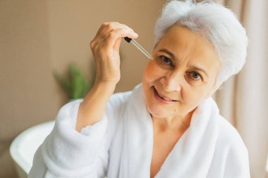 Cilde doğal damla kozmetik yağı süren bir kadın. Yetişkin yaşlı bir kadın günlük güzellik rutini yapan pipetli anti-yaş serumu uyguluyor. Yaşlı kadın yüz bakımı yaptırıyor.