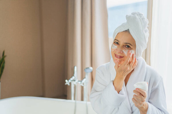 Mature adult old woman applying nourishing anti age face cream doing daily beauty routine. Senior woman with white mask or creme on skin gets facial treatment Skin care aging beauty skincare cosmetics