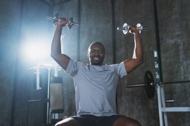 Beden eğitiminde spor. Spor salonunda dambılları toplayan Afrikalı Amerikalı bir vücut geliştirici. Fitness kulübünde halterli kasları çalıştırmak için ağırlık çalışıyorum. Spor yapan bir adam. Sağlıklı yaşam tarzı