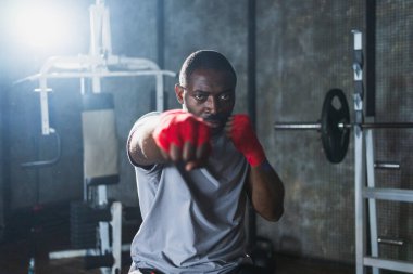 Beden eğitiminde spor. Afrikalı dövüşçü dövüşmeye hazırlanıyor kırmızı boksla yumruk atarken spor sargılarını spor salonunda sarar. Güçlü adam boks antrenmanına hazır yumrukları eğitiyor.