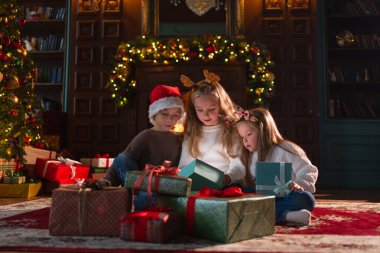 Mutlu Noeller Noel tatili. Çocuklar Noel ağacının yanındaki hediye kutusunu açıyor. Oturma odasında Noel ağacı şöminesi olan çocuklar sürpriz yüzlü hediye kutusunu açıyorlar. Evde Noel arifesi