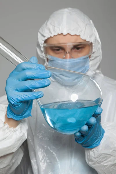 Scientific virologist. Biologist in protective suit and blue gloves holds big flask with blue substance sample. Flask with viruses concept. Virus University employee.