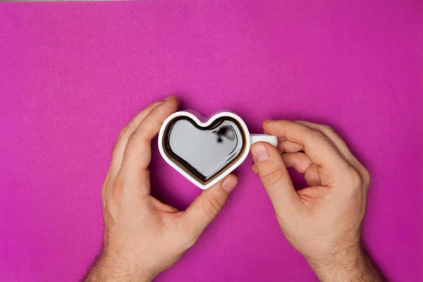 Man hands holds heart shaped cup of coffee on purple background. Black hot coffee in a white heart-shaped cup.