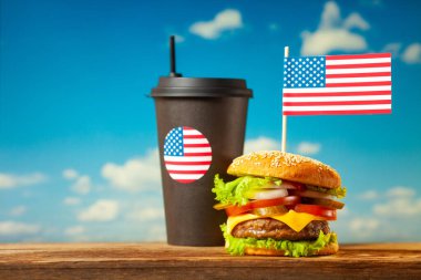 Close-up home made beef burger with american flag on the top and black paper cup on wooden table over blue sky background.