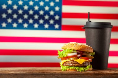 Close-up home made burger and black paper cup on wooden table over USA flag background.