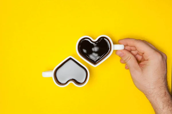 Man hands holds heart shaped cup of coffee on yellow background. Black hot coffee in a white heart-shaped cup. Flat lay.
