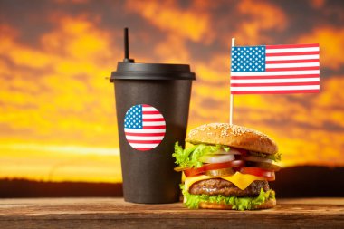 Yakın plan ev yapımı sığır eti burgeri, üstünde Amerikan bayrağı, üstünde de ahşap masa üzerinde saman olan siyah kağıt bardak..