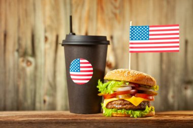 Close-up home made beef burger with american flag on the top and black paper cup with straw on wooden table over wooden background.