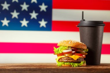 Close-up home made burger and black paper cup on wooden table over USA flag background.