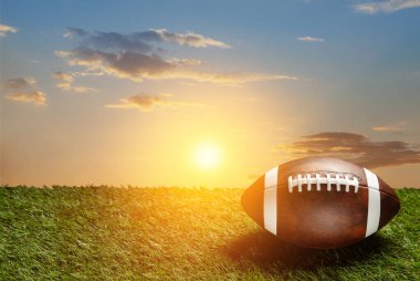 American football ball on green grass field on background of sunset sky. Banner with space for text.