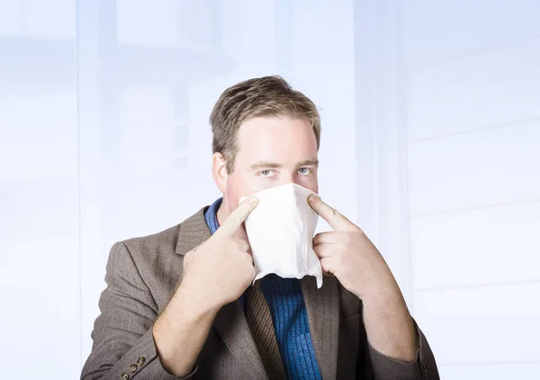 Unwell male office worker suffering from a contagious virus, covering face with tissue during cold and flu season