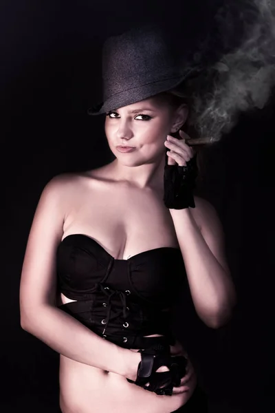 Stylish elegant woman in classy attire posing with cigar and hat on dark studio background in a smoking hot fashion concept