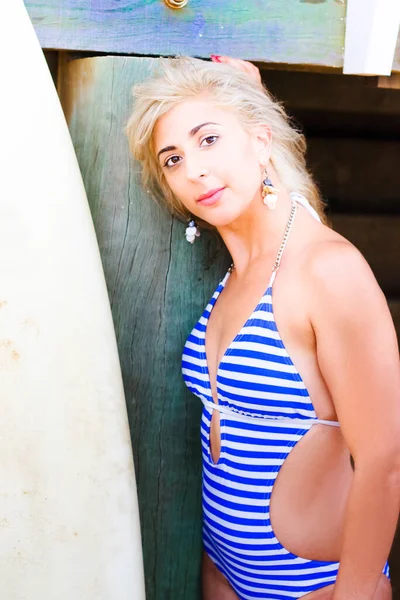 Pretty Blond Girl In Striped Swim Wear Sanding Next To A Beach Marina Pillar With Surf Board In A Healthy Summer Fitness Activity