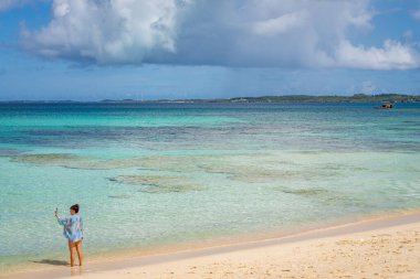 Okinawa sahilinde selfie çeken kadın turkuaz suyu ve güneşli gökyüzünü yakalıyor..