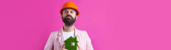 年轻的建筑工程师头戴橙色硬礼帽 穿着粉色夹克 站在红色工作室的背景 一个人拥有一座绿色的生态房子 — 图库照片