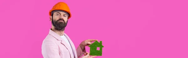 年轻的建筑工程师头戴橙色硬礼帽 穿着粉色夹克 站在红色工作室的背景 一个人拥有一座绿色的生态房子 — 图库照片