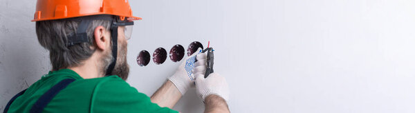 An electrician installs sockets in the apartment. A guy in an orange helmet and overalls makes electrics in the house.
