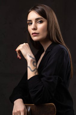 Stüdyoda uzun saçlı genç bir esmerin portresi. Koyu renkli dramatik bir fotoğraf. Kolunda yılan dövmesi olan bir kız..