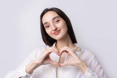 Gülümseyen genç bir kadının portresi beyaz arka planda parmakları izole bir şekilde kalp hareketi gösteriyor.