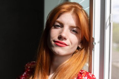 Kırmızı elbiseli, yüzünde bir gülümseme olan kızıl saçlı, aktif bir genç kız. Pencerenin yanında klasik bir portre.