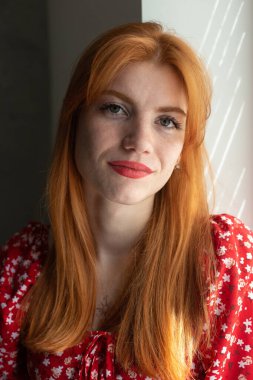 Kırmızı elbiseli, yüzünde bir gülümseme olan kızıl saçlı, aktif bir genç kız. Pencerenin yanında klasik bir portre.