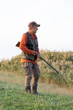 Olgun bir avcı elinde pompalı tüfekle tarlada yürüyor.