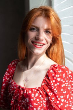 Kırmızı elbiseli, yüzünde bir gülümseme olan kızıl saçlı, aktif bir genç kız. Pencerenin yanında klasik bir portre.