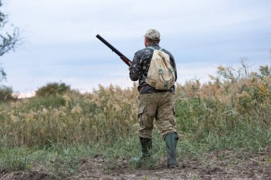 Olgun bir avcı elinde pompalı tüfekle tarlada yürüyor.