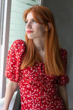 Kırmızı elbiseli, yüzünde bir gülümseme olan kızıl saçlı, aktif bir genç kız. Pencerenin yanında klasik bir portre.