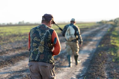 Olgun bir avcı elinde pompalı tüfekle tarlada yürüyor.