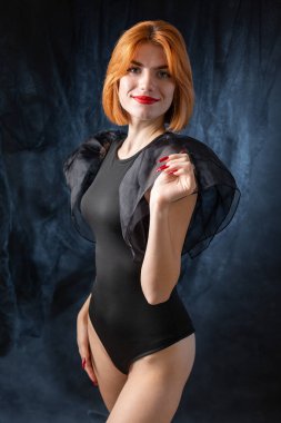 Siyah elbiseli genç, kızıl saçlı bir kız stüdyoda koyu bir arka plana karşı poz veriyor..