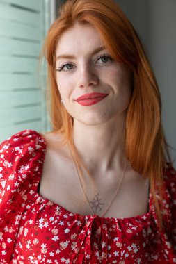 Kırmızı elbiseli, yüzünde bir gülümseme olan kızıl saçlı, aktif bir genç kız. Pencerenin yanında klasik bir portre.