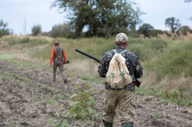 Olgun bir avcı elinde pompalı tüfekle tarlada yürüyor.
