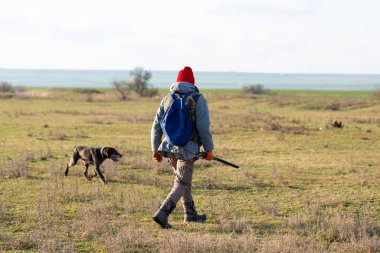 Olgun bir avcı elinde pompalı tüfekle tarlada yürüyor.