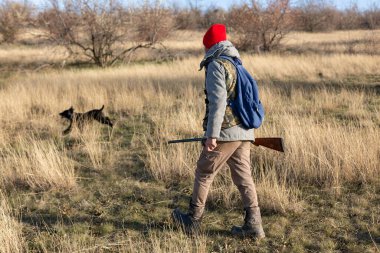 Olgun bir avcı elinde pompalı tüfekle tarlada yürüyor.