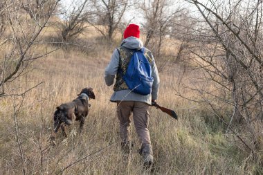 Olgun bir avcı elinde pompalı tüfekle tarlada yürüyor.