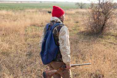 Olgun bir avcı elinde pompalı tüfekle tarlada yürüyor.