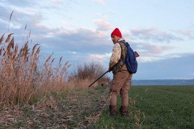 Olgun bir avcı elinde pompalı tüfekle tarlada yürüyor.