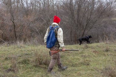 Olgun bir avcı elinde pompalı tüfekle tarlada yürüyor.