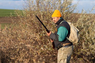 Olgun bir avcı elinde pompalı tüfekle tarlada yürüyor.