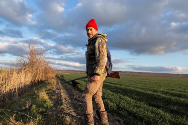 Olgun bir avcı elinde pompalı tüfekle tarlada yürüyor.
