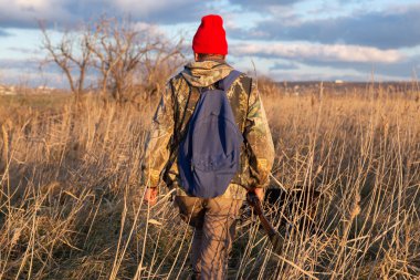 Olgun bir avcı elinde pompalı tüfekle tarlada yürüyor.