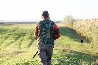 Olgun bir avcı elinde pompalı tüfekle tarlada yürüyor.