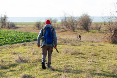 Olgun bir avcı elinde pompalı tüfekle tarlada yürüyor.