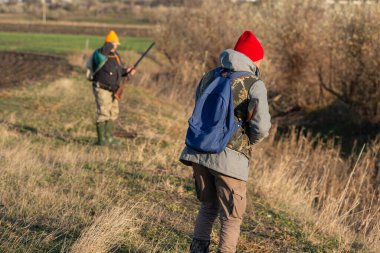 Olgun bir avcı elinde pompalı tüfekle tarlada yürüyor.