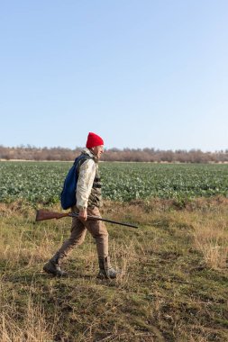 Olgun bir avcı elinde pompalı tüfekle tarlada yürüyor.