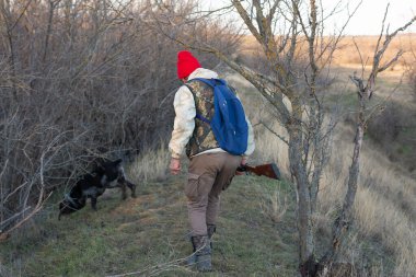 Olgun bir avcı elinde pompalı tüfekle tarlada yürüyor.