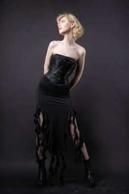 Stüdyoda bir şilin altında etkileyici genç bir sarışın. Siyah bir korse ve elbiseyle.