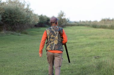 Olgun bir avcı elinde pompalı tüfekle tarlada yürüyor.